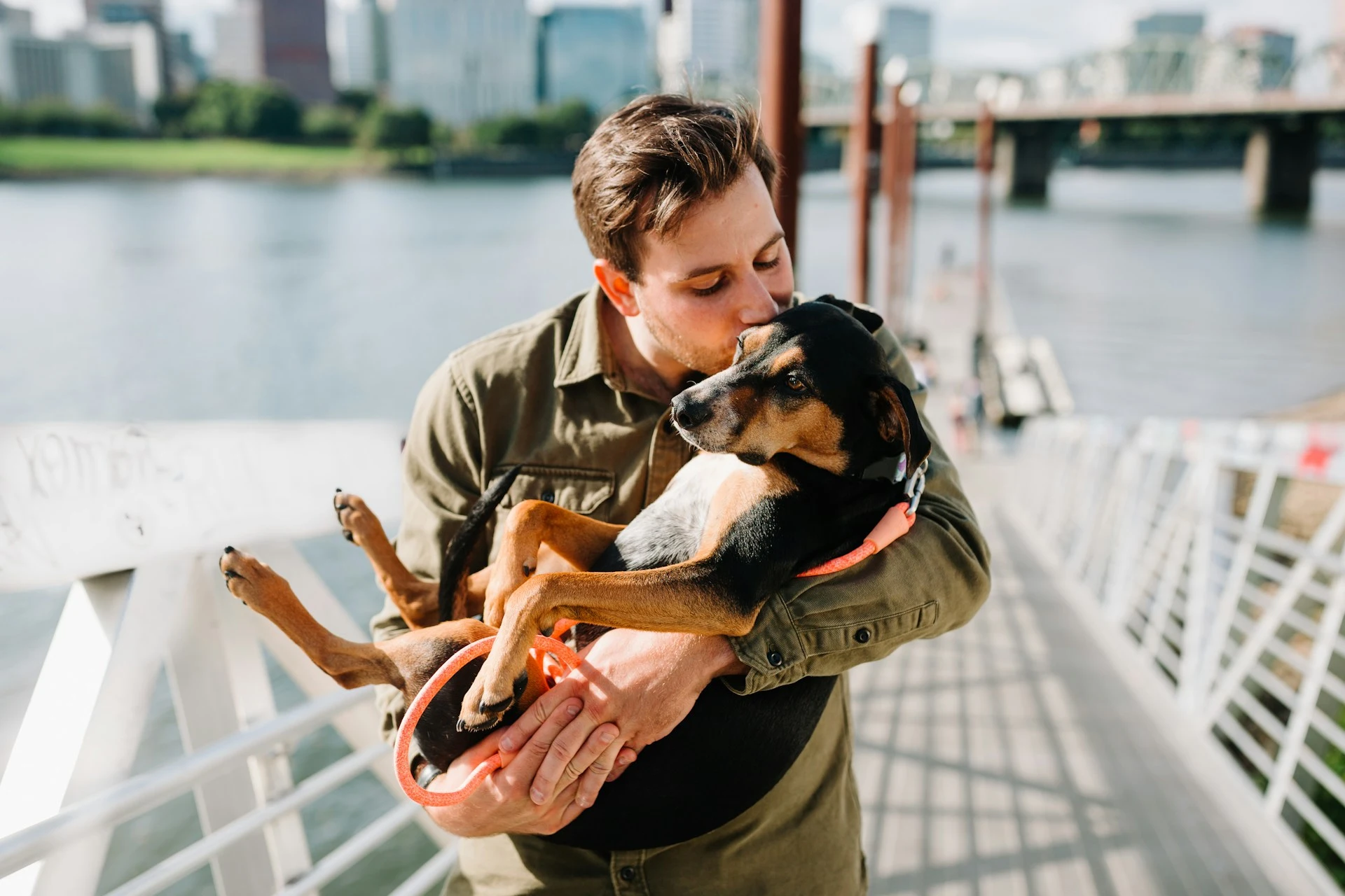A man kisses the top of his dog's head while cradling it in his arms on a sunny bridge overlooking a river and city skyline.